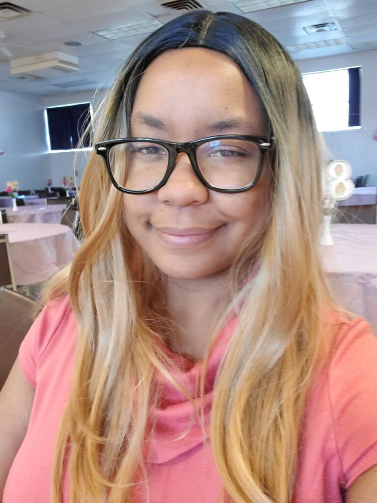 Photo of Angel, a young Black genderfluid person with long light-colored straightened hair, smiles at the camera. They are in a conference room with many tables set up, and they are wearing a pink cowled collar shirt.