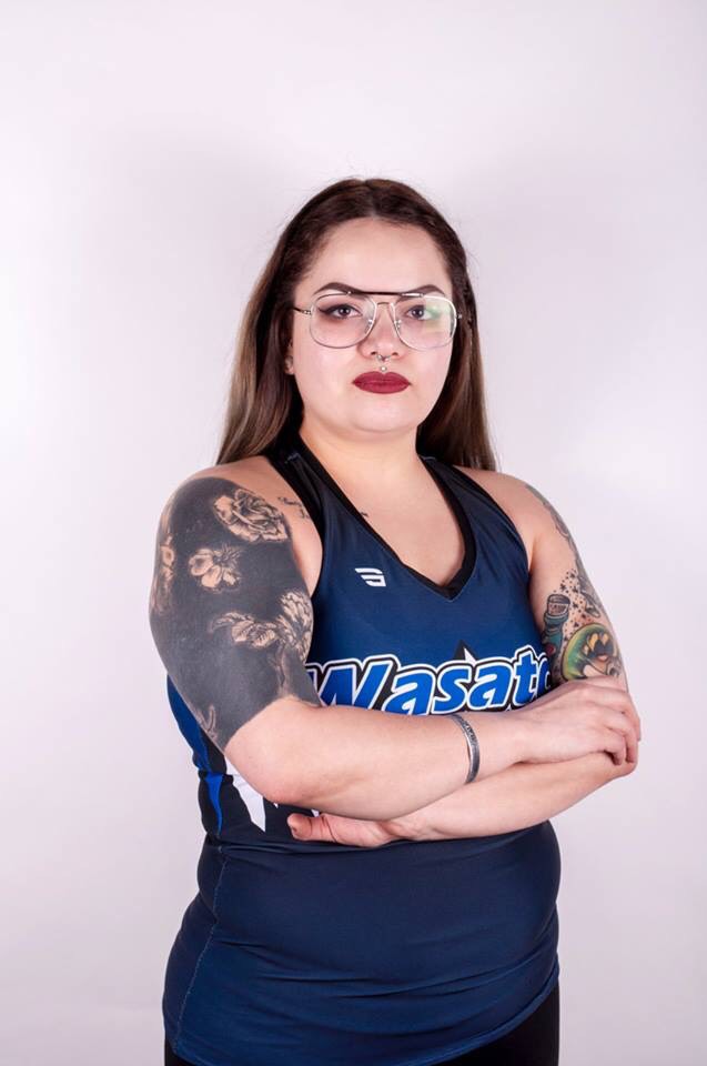 Photo of Brianna, a young Latinx person with glasses and highlights in their straight dark hair, looks directly at the camera, crossing their arms. She is wearing a tank top over black pants, and has half-sleeve tattoos on both of her arms, as well as part of a tattoo of cursive script on part of her chest. They are wearing a silver bracelet and crimson lipstick.