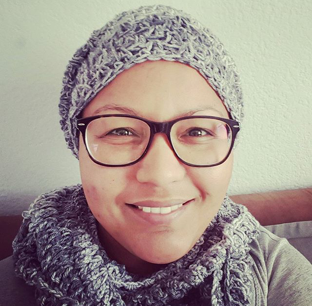 Photo of Chevone, a young Cape Coloured woman, smiling at the camera. She is wearing a knit silver hat and matching scarf over a grey shirt, and glasses.