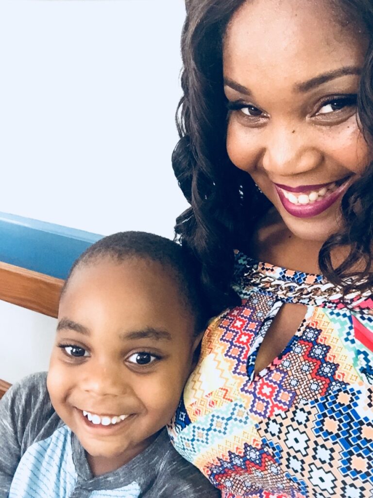 Photo of Shamiir, a young Caribbean-descended person of color with short hair, smiling with his mother Nikita, who has wavy shoulder-length hair. Shamiir is wearing a green and grey t-shirt, and Nikita is wearing a bright multi-colored woven-fabric pattern shirt.