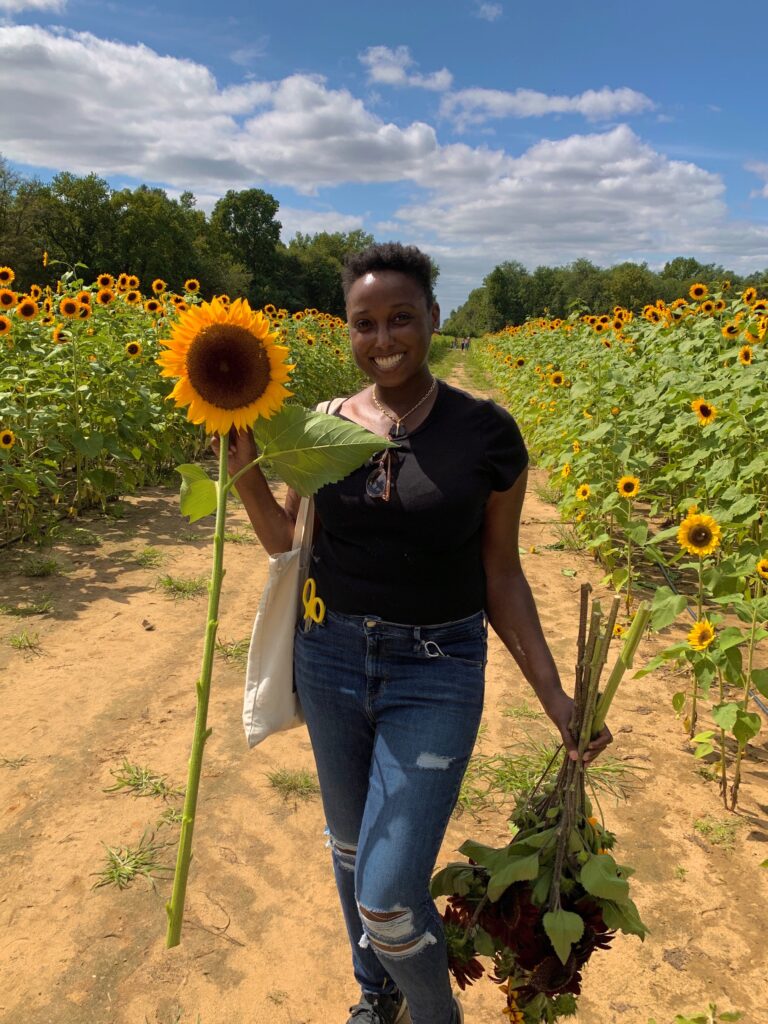 A Black person standing in the middle of a sunflower patch, holding a very tall sunflower in one hand and a bunch of various colored sunflowers in the other, wearing a black shirt and blue ripped jeans, smiling, with short hair.