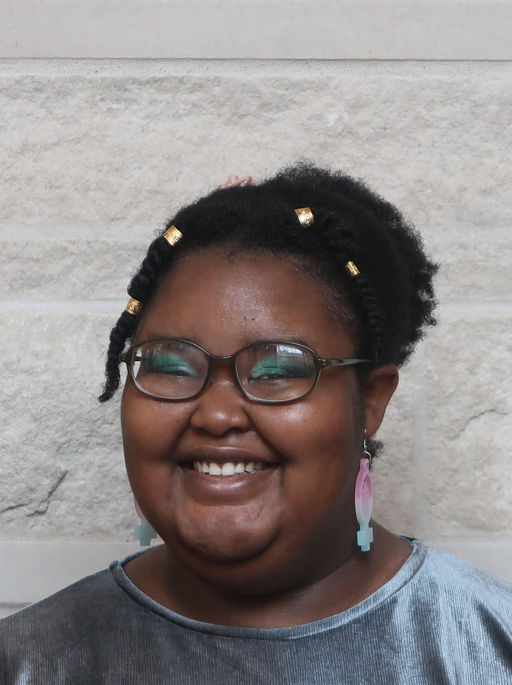 A smiling Black individual is pictured against a white, stone background. Their head is tilted to the left. They are wearing a silver blouse and translucent brown glasses, and have their afro in a bun with two twists framing their face. They have on shimmery green, ombré eyeshadow and are wearing feminist fist earrings in the trans flag’s colors.