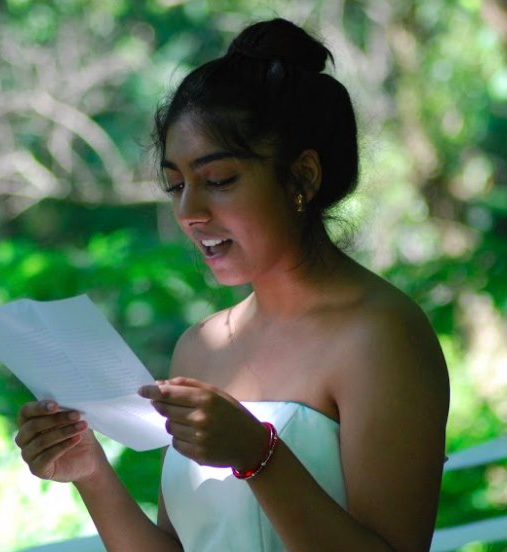 A young Indian woman in a white strapless dress with her hair up in a bun stands in front of a backdrop of trees. She smiles, looking down at a paper from which she is reading aloud.