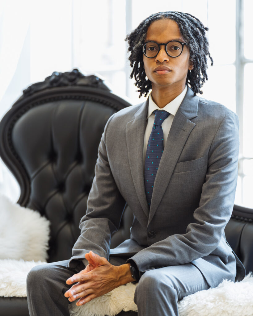 A young, Black man with short loc'd hair almost reaching his shoulders. He has a stern facial expression. He's wearing: black, square glasses; a light, gray suit; a white shirt; and a navy blue tie that has light blue dots all over the tie. He's sitting in a room with plenty of natural light and he's sitting on a white, faux fur blanket that's laid across a black leather sofa.