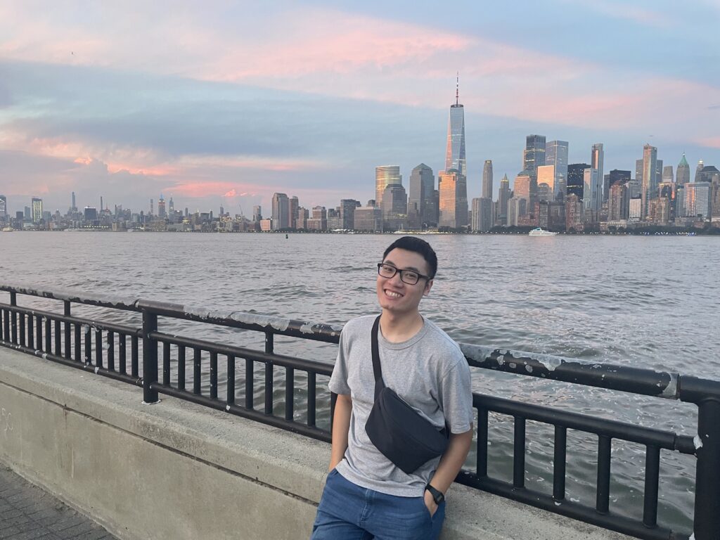 A young Vietnamese man in a grey t-shirt and his jeans. Both of his hands are in pockets. He is leaning on the railing of the Hudson River during the sunset, with many tall buildings of Manhattan behind him. His head is tilted to the left and he is smiling gently to the camera.