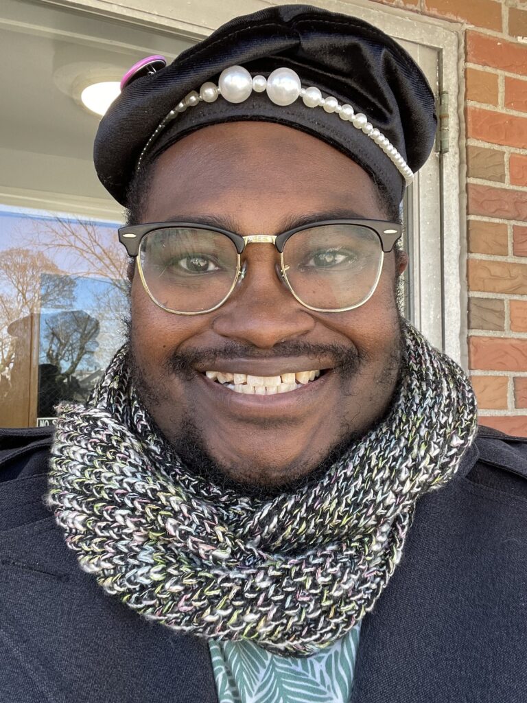 A photo of a smiling Black man in his midthirties. He’s wearing black-and-gold glasses, a floppy velvet hat covered with pins, a black coat, a t-shirt printed with green leaves on it, and a gold infinity scarf around his neck.