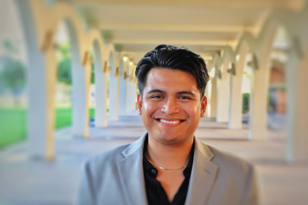 Photo of Ariel, a person of Mexican descent, wearing a black collared shirt and grey blazer, smiling outside between a row of high-arched columns.